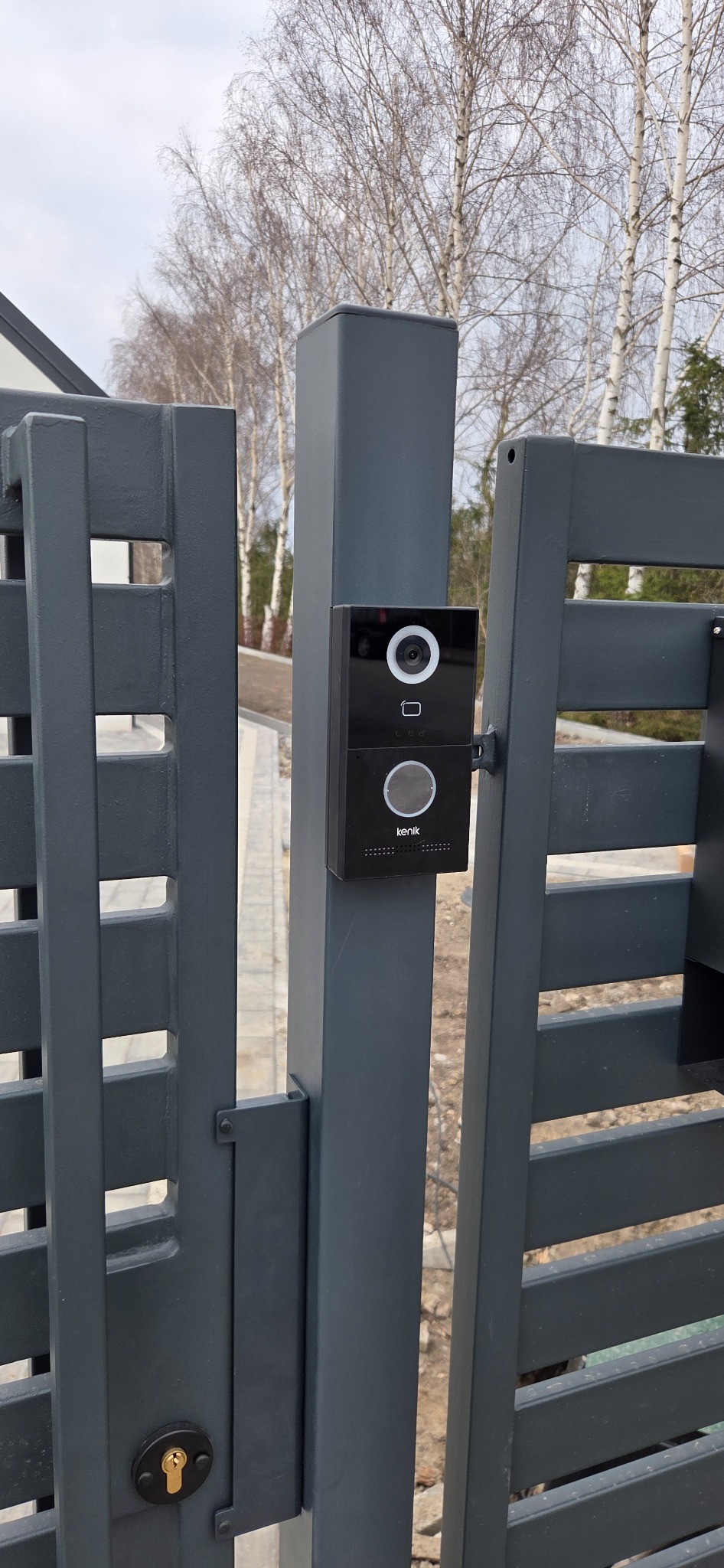 Nowoczesny wideodomofon Kenik zamontowany na grafitowej bramie posesji. Widoczne drzewa w tle. Minimalistyczny design urządzenia.