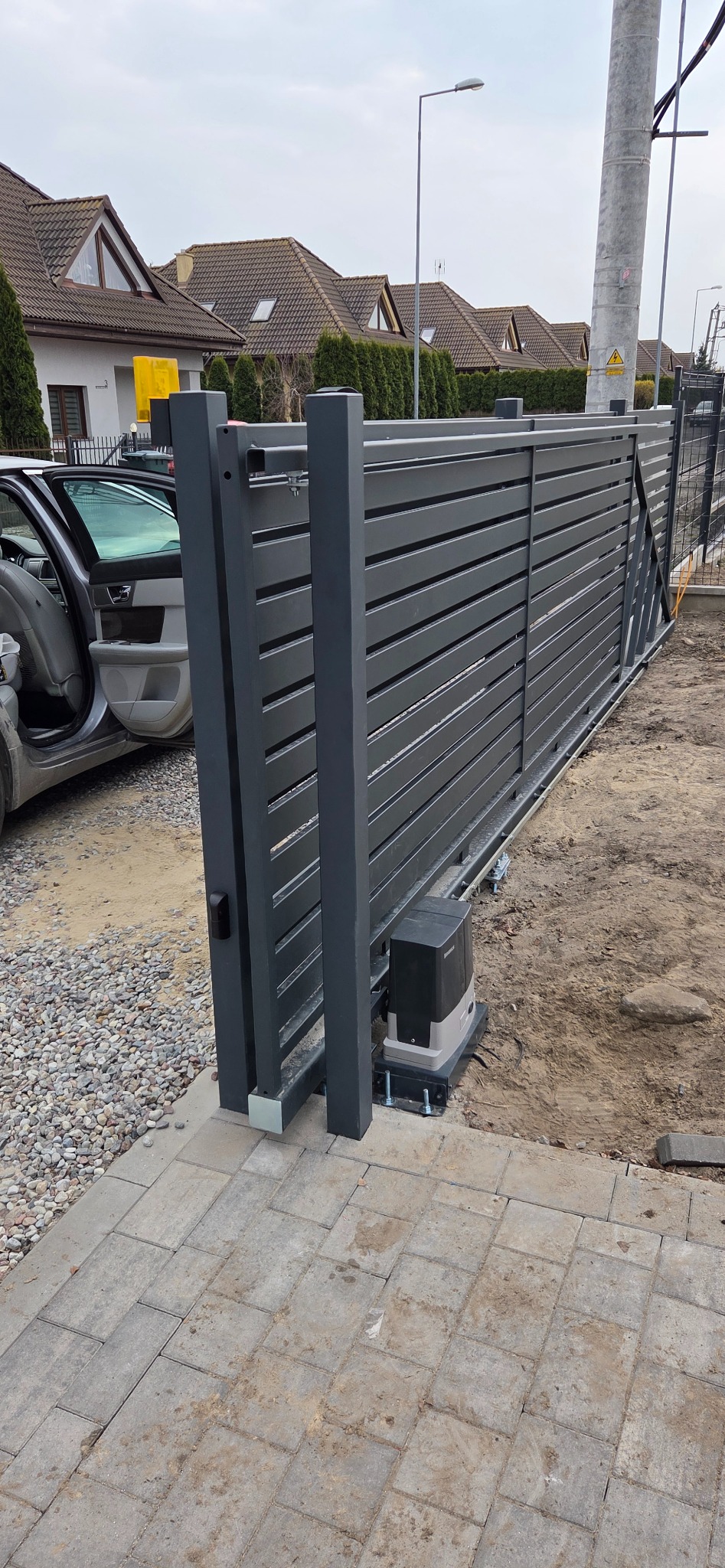 Nowoczesna, przesuwna brama posesyjna z automatyką, widoczna z boku. Szara, pozioma konstrukcja z napędem. W tle domy jednorodzinne i częściowo otwarty samochód.