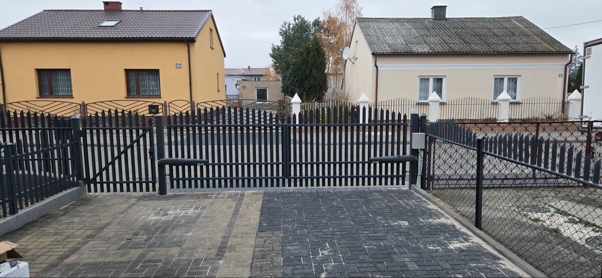Nowoczesna, czarna brama automatyczna z napędem ramieniowym, zamontowana przed domem z kostki brukowej. Widoczne dwa budynki w tle, jeden w odcieniach żółci, drugi beżowy.