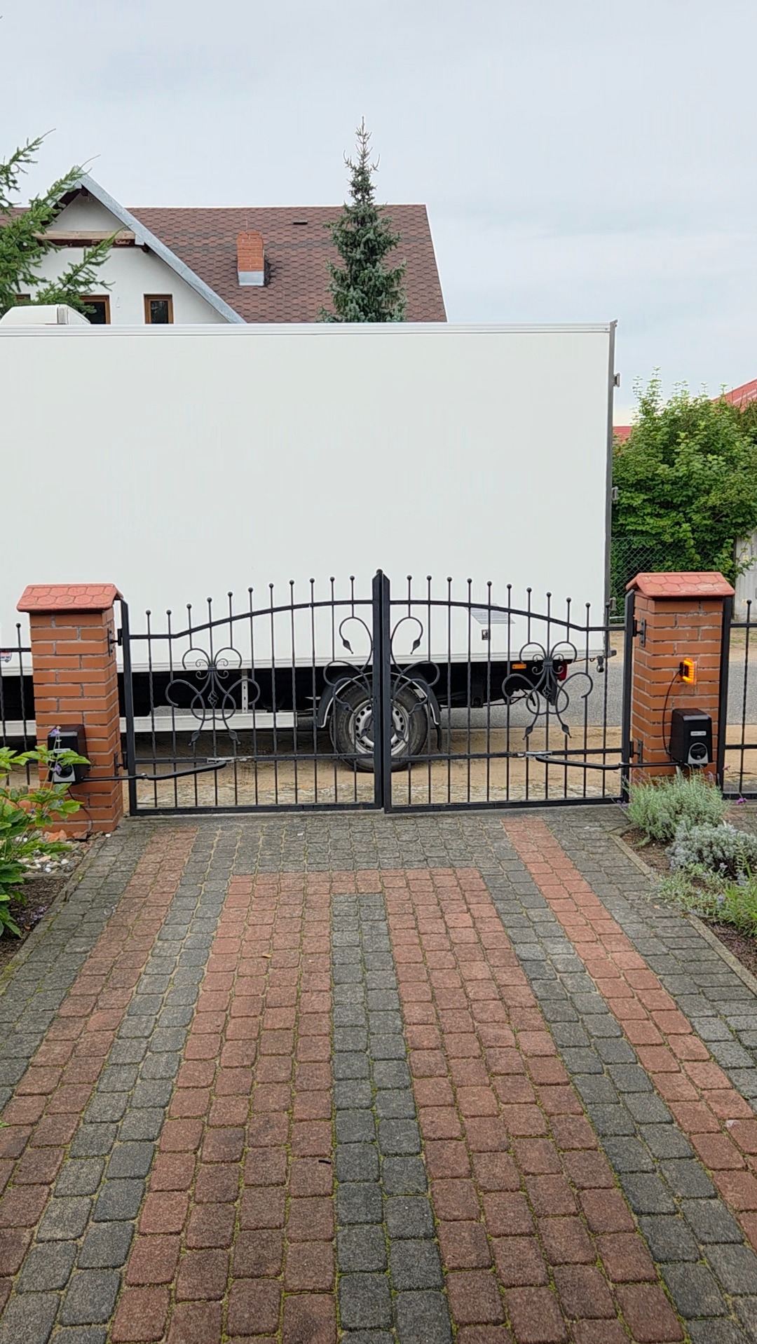 Kuta brama wjazdowa z automatyką, czarna, z dekoracyjnymi elementami, zamontowana na ceglanych słupkach. W tle biały samochód dostawczy i dom z brązowym dachem.