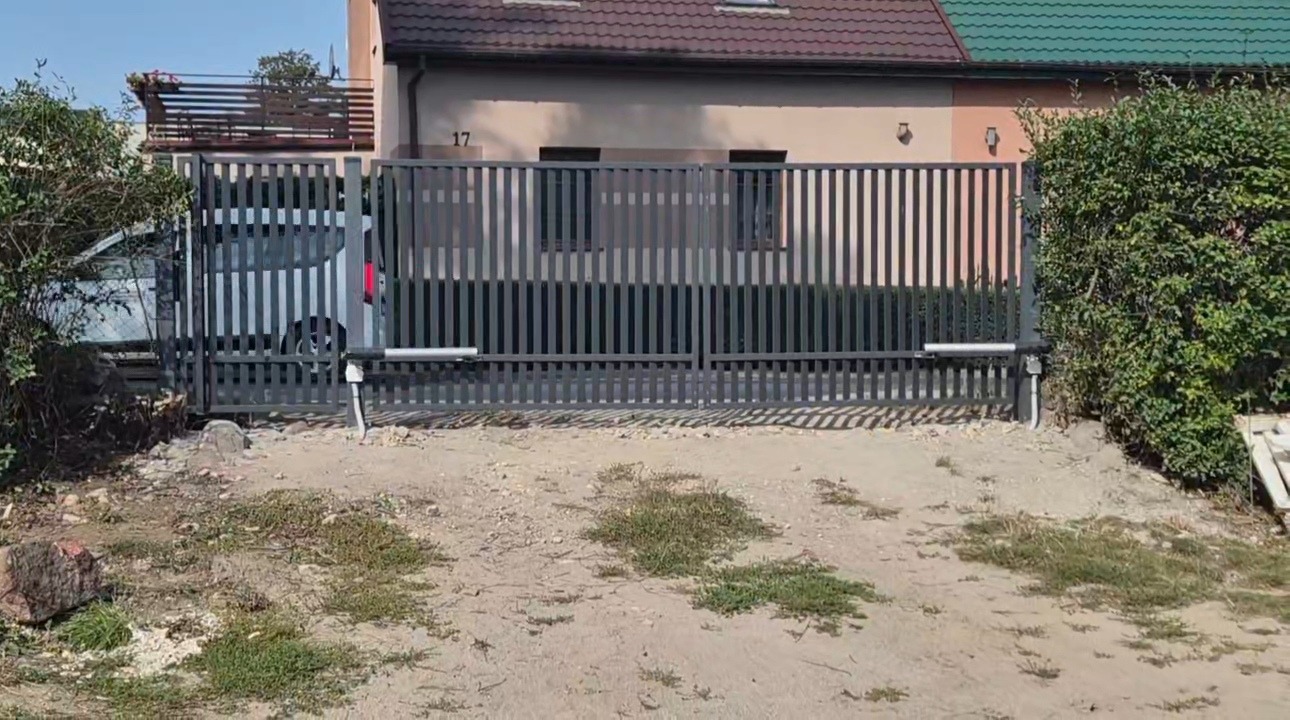 Szara, przesuwna brama z automatyką na tle domu z numerem 17. Widoczny samochód za bramą i zielona roślinność po bokach. Brama z pionowych, metalowych elementów.