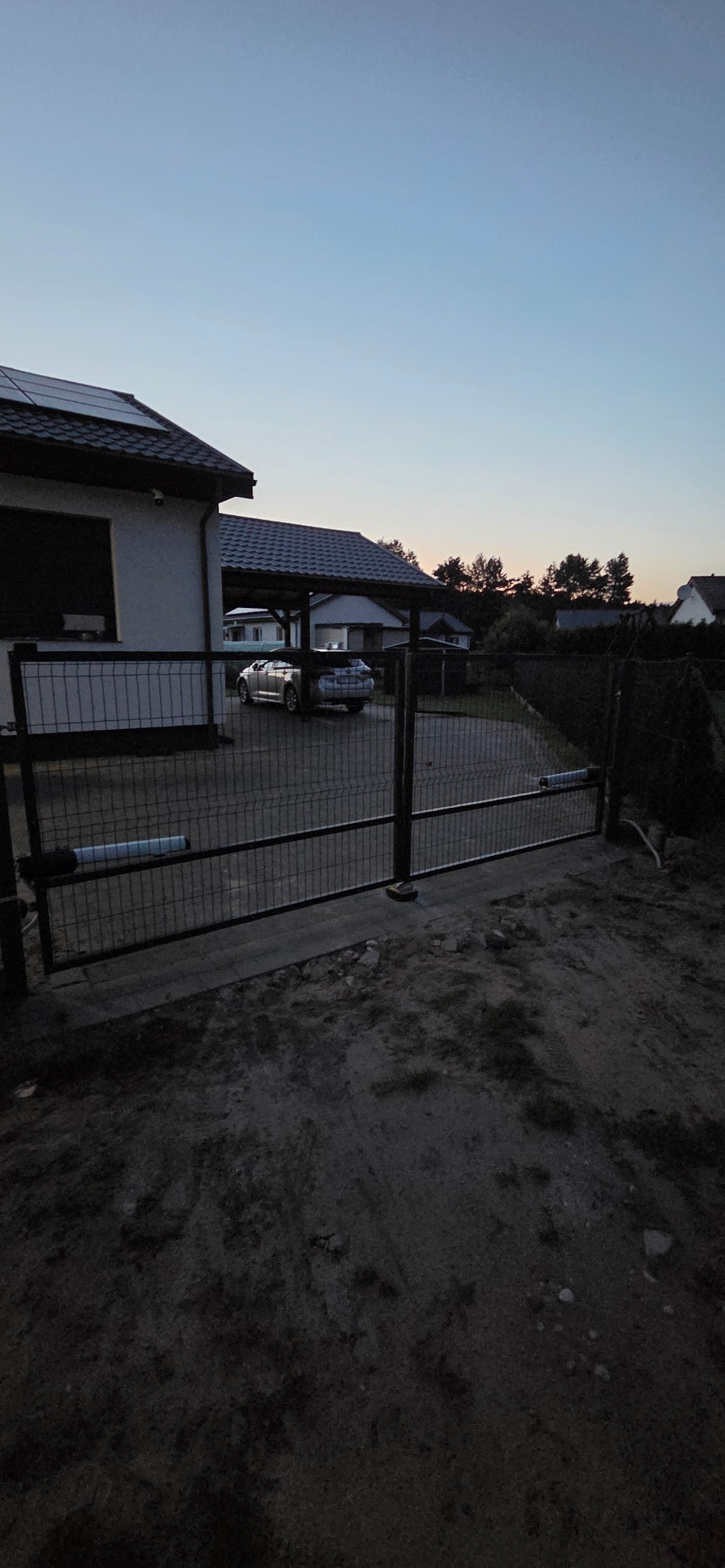 Nowoczesna brama posesyjna z automatyką, widoczna na tle domu z panelami słonecznymi i zadaszeniem dla samochodów o zmierzchu. Minimalistyczny design.