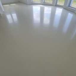 Karol Zajączkowski - Jasna, jednolita podłoga z połyskiem w przestronnym wnętrzu z kominkiem i oknami. Gładka powierzchnia odbija światło, tworząc wrażenie czystości i nowoczesności.