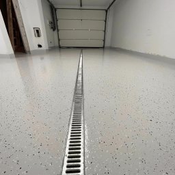 Karol Zajączkowski - Jasnoszara posadzka z odpływem liniowym w garażu. Widoczna brama garażowa i fragment ściany. Posadzka z delikatnym wzorem w czarne i białe plamki.