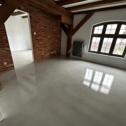 Karol Zajączkowski - Jasnoszara, gładka posadzka w loftowym wnętrzu z ceglaną ścianą i drewnianymi belkami stropowymi. Okno z ciemną ramą odbija się w powierzchni podłogi.