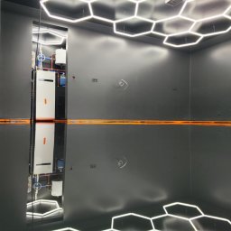 Karol Zajączkowski - Błyszcząca, ciemna posadzka z odbiciem heksagonalnych lamp sufitowych. W tle widoczny system grzewczy, co sugeruje przemysłowy charakter wnętrza.