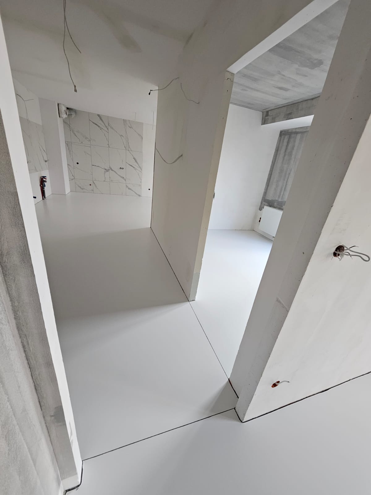 Biała, gładka posadzka żywiczna w surowym wnętrzu. Widoczne ściany w trakcie wykończenia, marmurowe płytki na ścianie w tle. Minimalistyczny design, surowy beton na suficie.