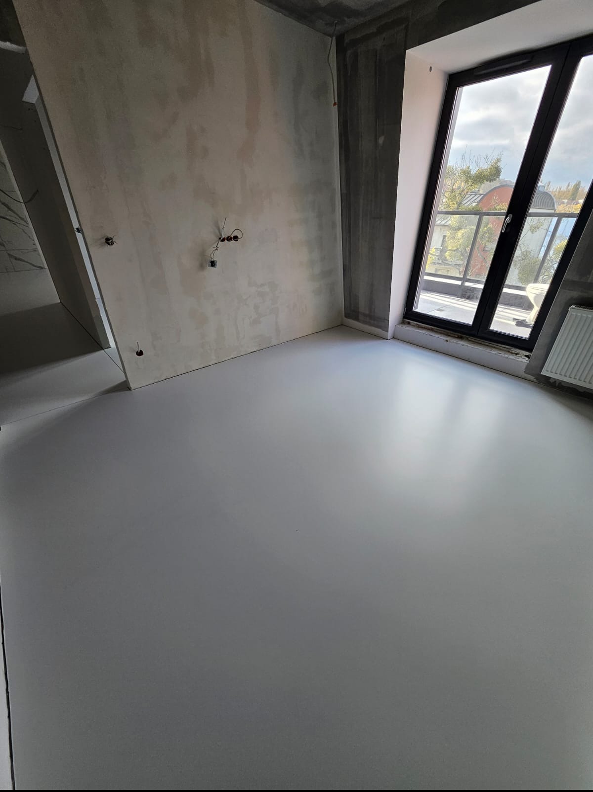 Jasnoszara, gładka podłoga w surowym wnętrzu z oknem balkonowym. Widoczne niedokończone ściany i instalacje elektryczne. Minimalistyczny design.