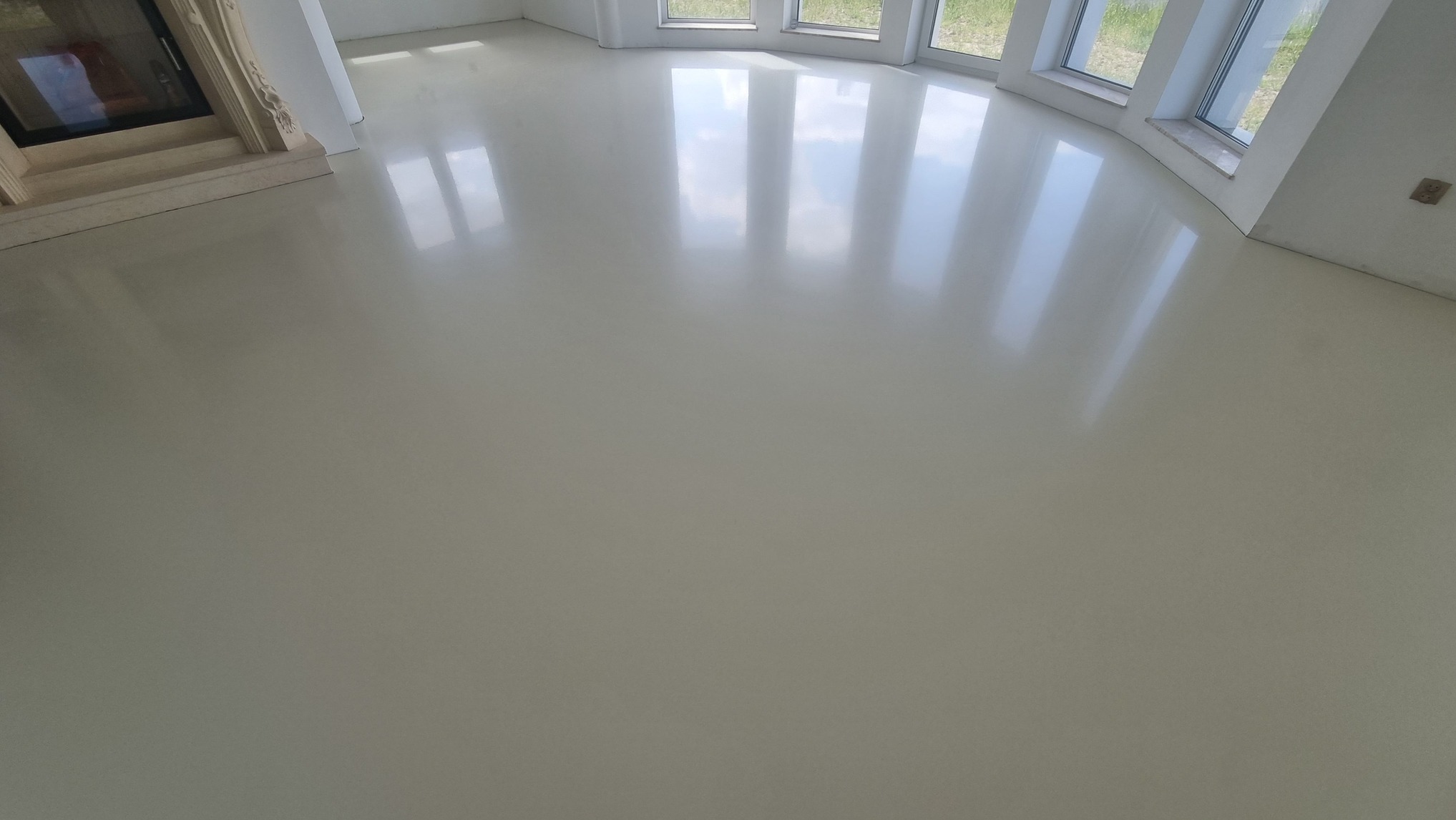 Jasna, jednolita podłoga z połyskiem w przestronnym wnętrzu z kominkiem i oknami. Gładka powierzchnia odbija światło, tworząc wrażenie czystości i nowoczesności.