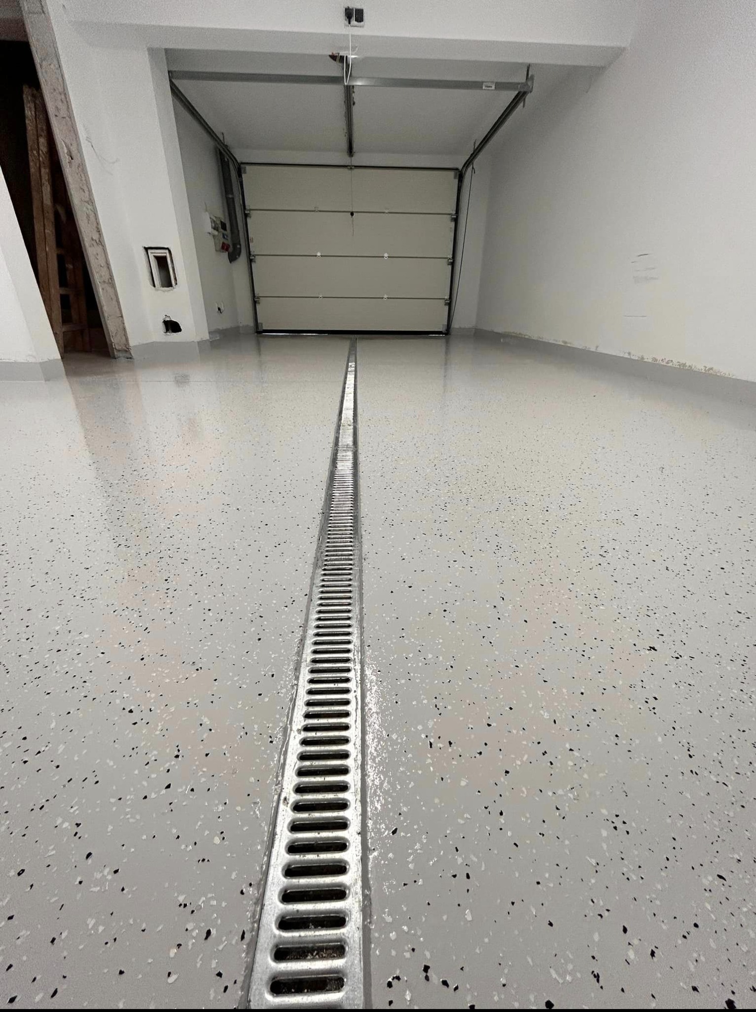 Jasnoszara posadzka z odpływem liniowym w garażu. Widoczna brama garażowa i fragment ściany. Posadzka z delikatnym wzorem w czarne i białe plamki.
