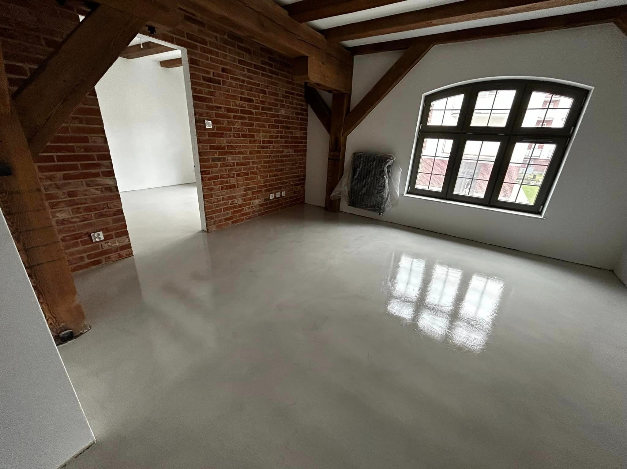 Jasnoszara, gładka posadzka w loftowym wnętrzu z ceglaną ścianą i drewnianymi belkami stropowymi. Okno z ciemną ramą odbija się w powierzchni podłogi.