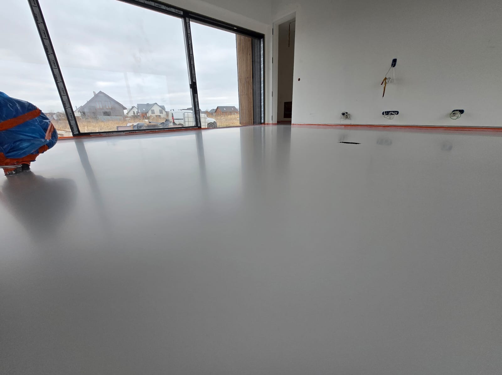 Jasnoszara, gładka posadzka w nowym wnętrzu z dużym oknem. Widoczne zabezpieczenia przy ścianach i częściowo zakryty sprzęt budowlany. Minimalistyczny design.