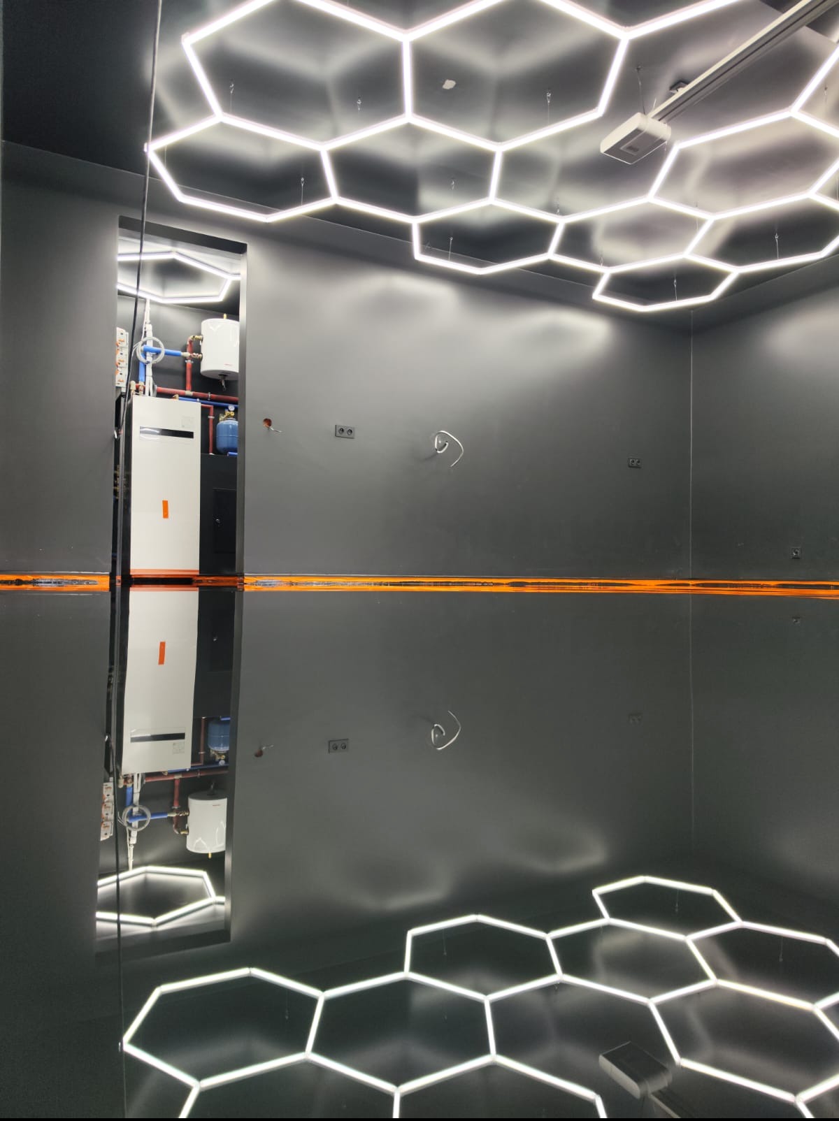 Błyszcząca, ciemna posadzka z odbiciem heksagonalnych lamp sufitowych. W tle widoczny system grzewczy, co sugeruje przemysłowy charakter wnętrza.