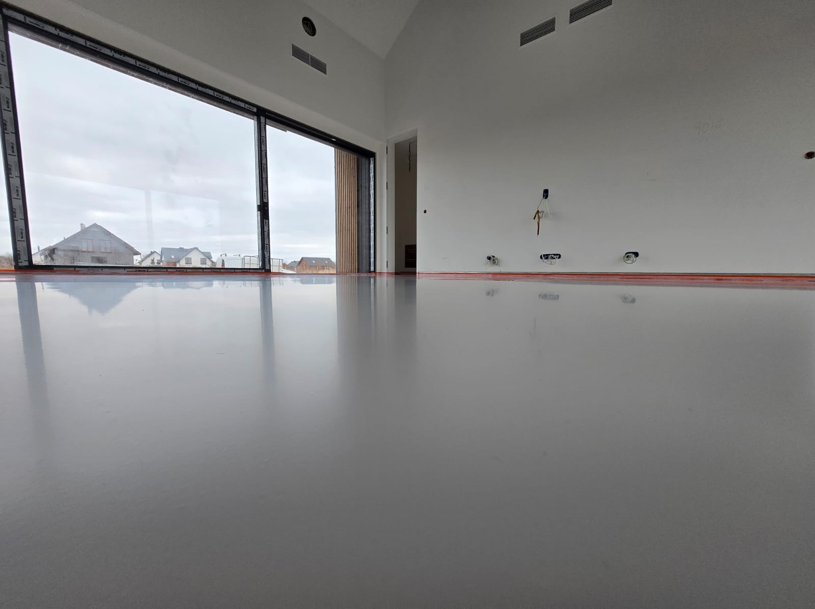 Błyszcząca, szara posadzka żywiczna w nowym domu z dużym oknem i widokiem na osiedle. Widoczne przygotowane punkty elektryczne na ścianie.