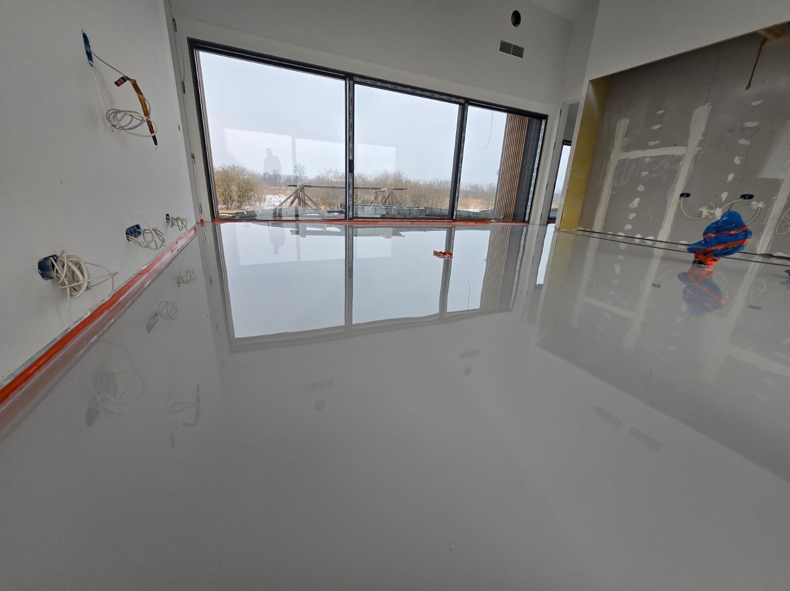 Błyszcząca, szara posadzka w jasnym wnętrzu z dużymi oknami. Widoczne niedokończone ściany i wystające kable elektryczne. Minimalistyczny design, gładka powierzchnia.