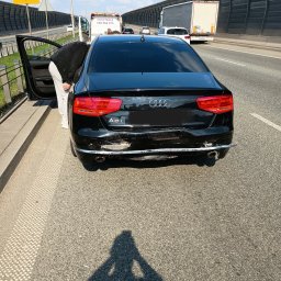 boomboom car - Uszkodzony tył czarnego Audi A8 na poboczu autostrady, otwarte drzwi, w tle laweta i znak ostrzegawczy. Widoczne wgniecenia i zarysowania na zderzaku.