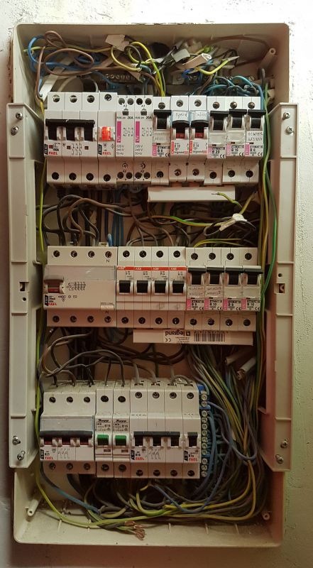 Otwarta skrzynka elektryczna z bezpiecznikami i przewodami wewnątrz, widok z góry. Różne typy bezpieczników, przewody w kolorach: niebieski, żółty, zielony.