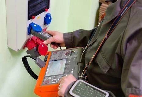 Elektryk w kombinezonie wykonuje pomiary elektryczne gniazd siłowych za pomocą specjalistycznego miernika z ekranem LCD i sondą pomiarową.