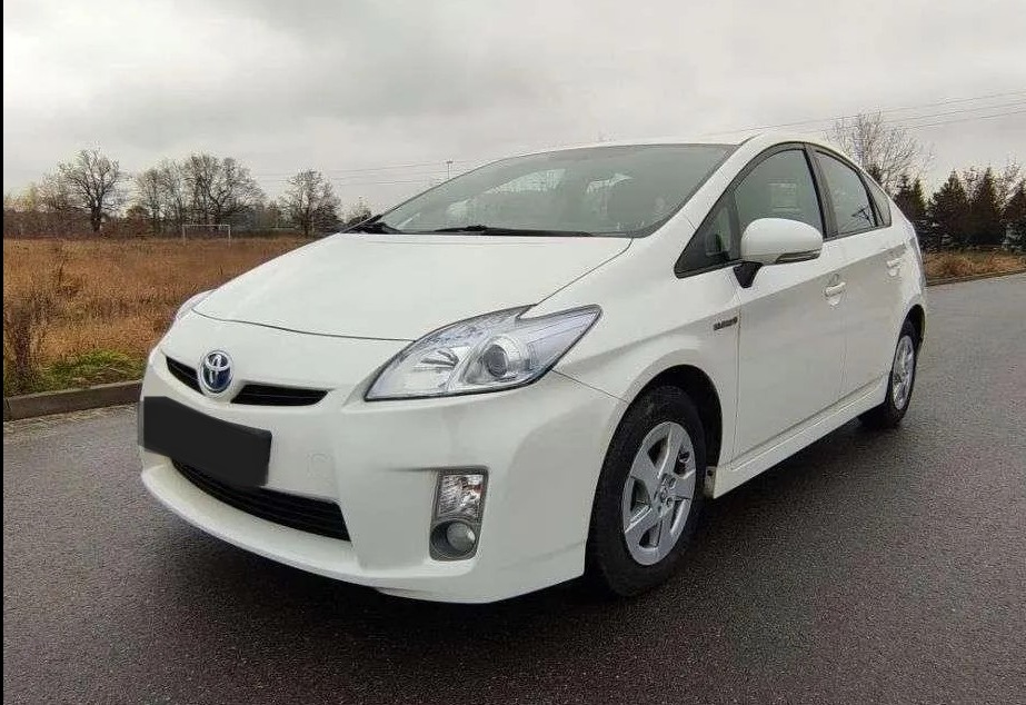 Biała Toyota Prius na zewnątrz, widok z przodu, lekko z boku. Auto stoi na mokrym asfalcie, w tle pole i drzewa pod pochmurnym niebem. Widoczny fragment boiska.