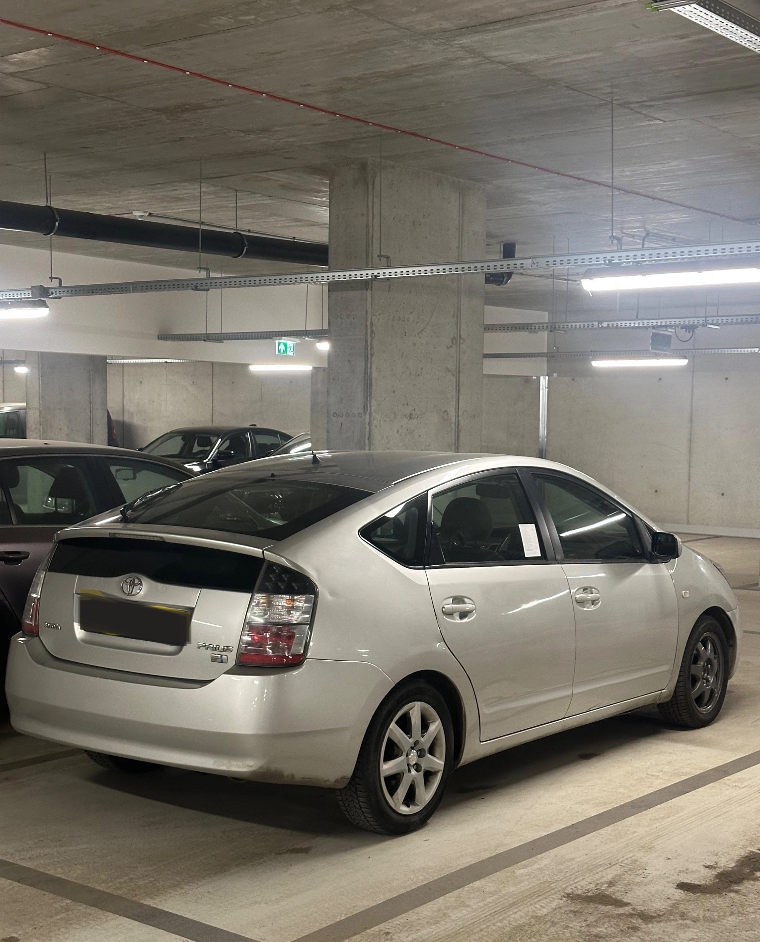 Srebrny Toyota Prius zaparkowany w garażu podziemnym, widok z tyłu. Widoczne betonowe filary i instalacje pod sufitem. Oświetlenie jarzeniowe.