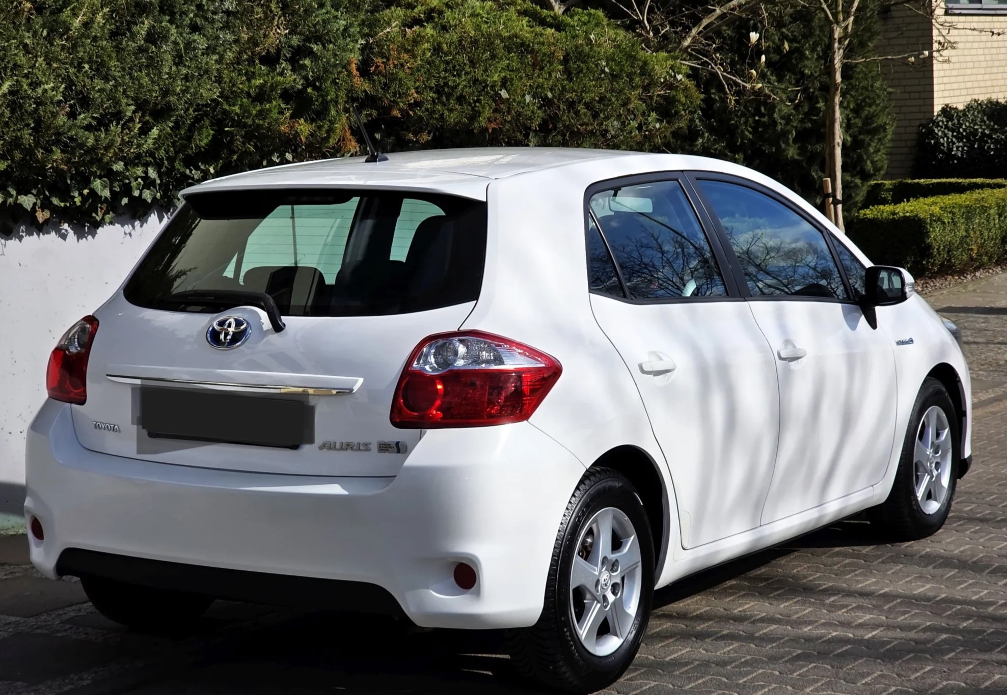 Biała Toyota Auris z tyłu, widoczne światła, logo i oznaczenie modelu. Auto stoi na kostce brukowej, w tle zieleń krzewów i drzew.