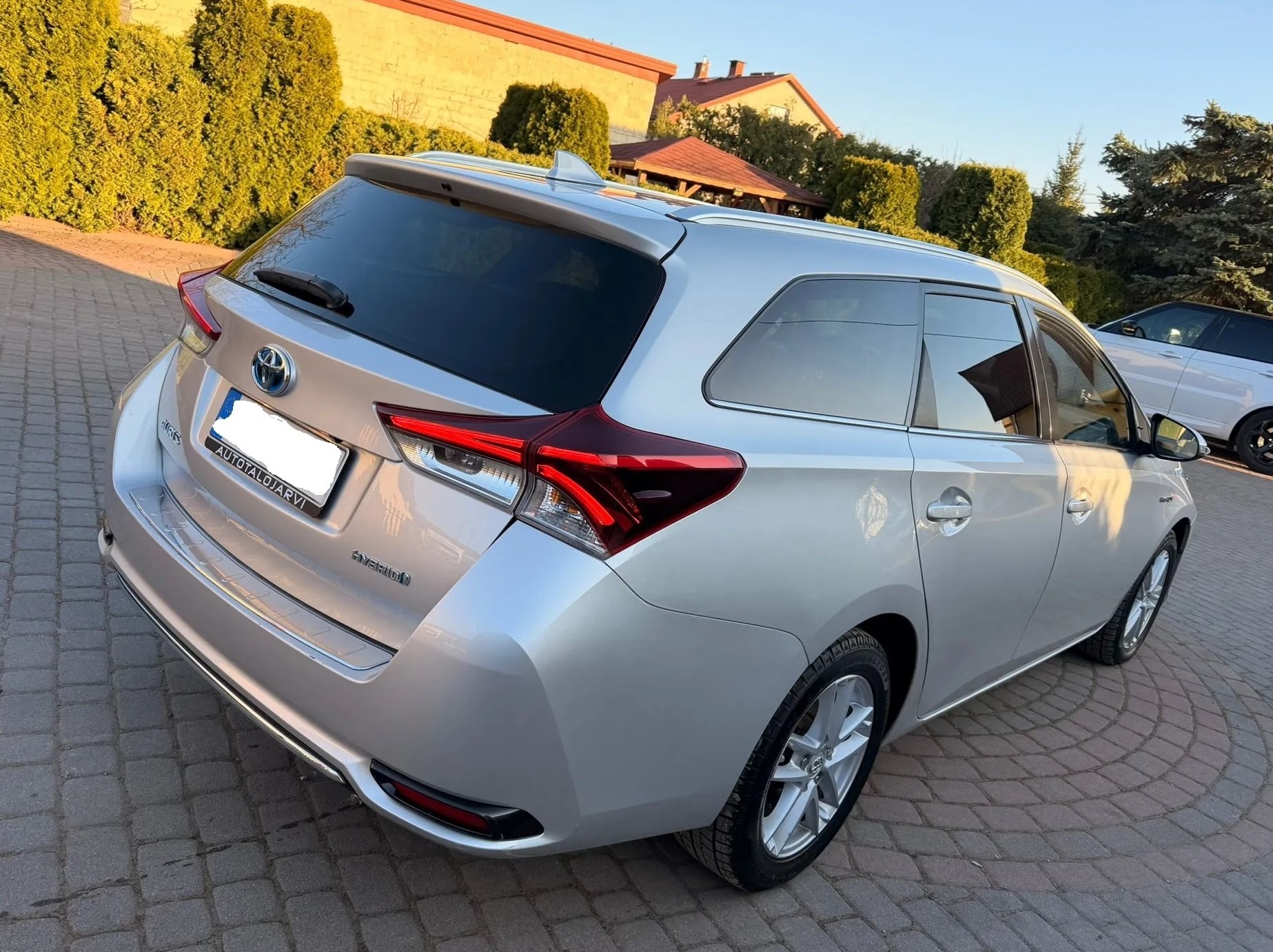 Srebrny samochód kombi Toyota Auris Hybrid, widok z tyłu, zaparkowany na kostce brukowej. W tle budynek z żółtej cegły i zielone krzewy.