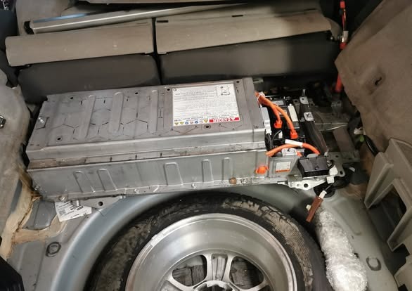 Akumulator hybrydowy w bagażniku samochodu, widoczne przewody i koło zapasowe. Metalowa obudowa akumulatora z naklejkami ostrzegawczymi.