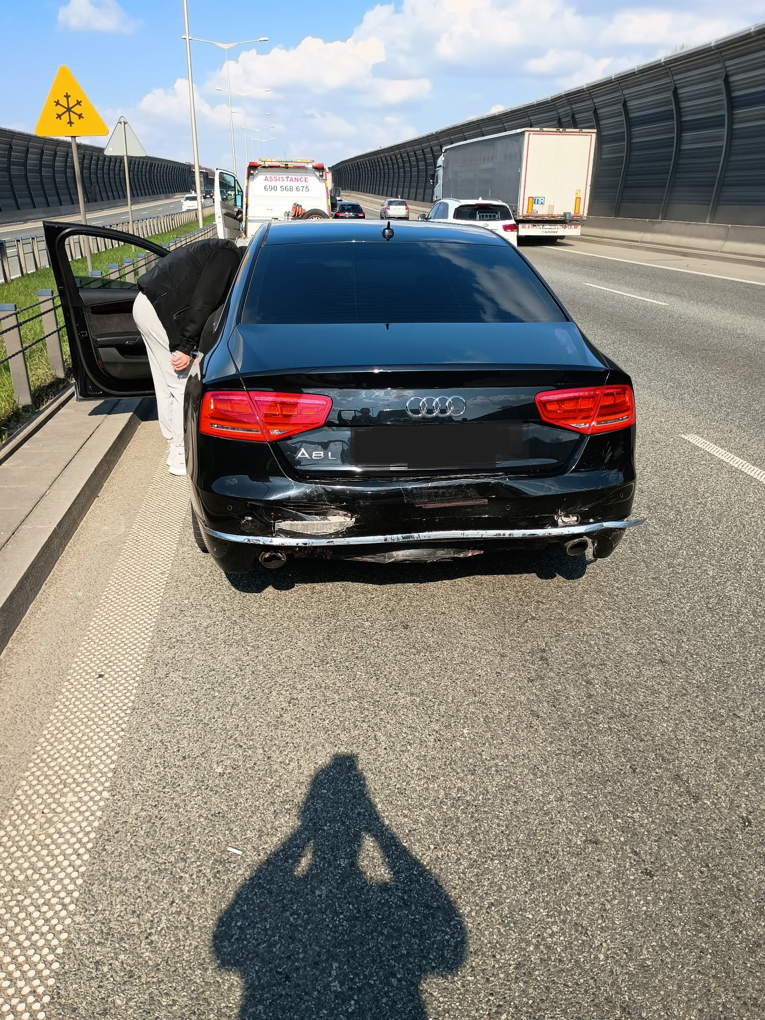 Uszkodzony tył czarnego Audi A8 na poboczu autostrady, otwarte drzwi, w tle laweta i znak ostrzegawczy. Widoczne wgniecenia i zarysowania na zderzaku.