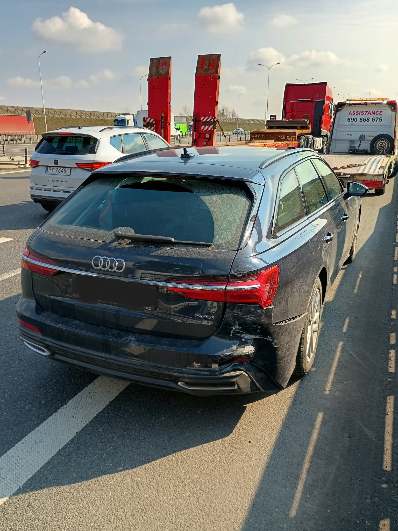 Uszkodzony tył czarnego Audi A6 Avant po kolizji, widoczny na poboczu drogi, z lawetą w tle. Zarysowania i wgniecenia na zderzaku i błotniku.