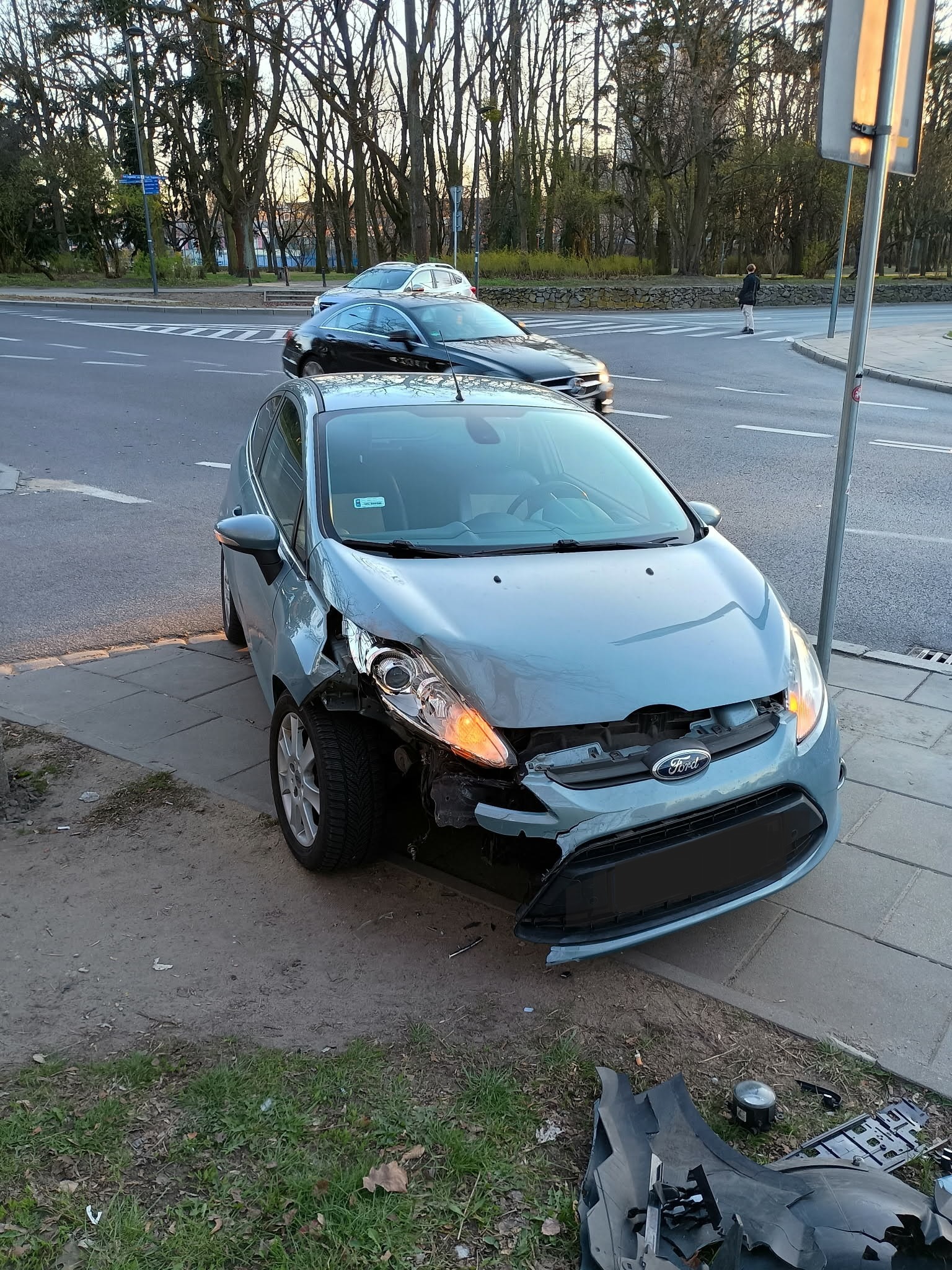 Uszkodzony, błękitny Ford Fiesta po kolizji, zaparkowany na chodniku. Widoczne wgniecenia na karoserii i brakujące elementy zderzaka. W tle ulica i drzewa.