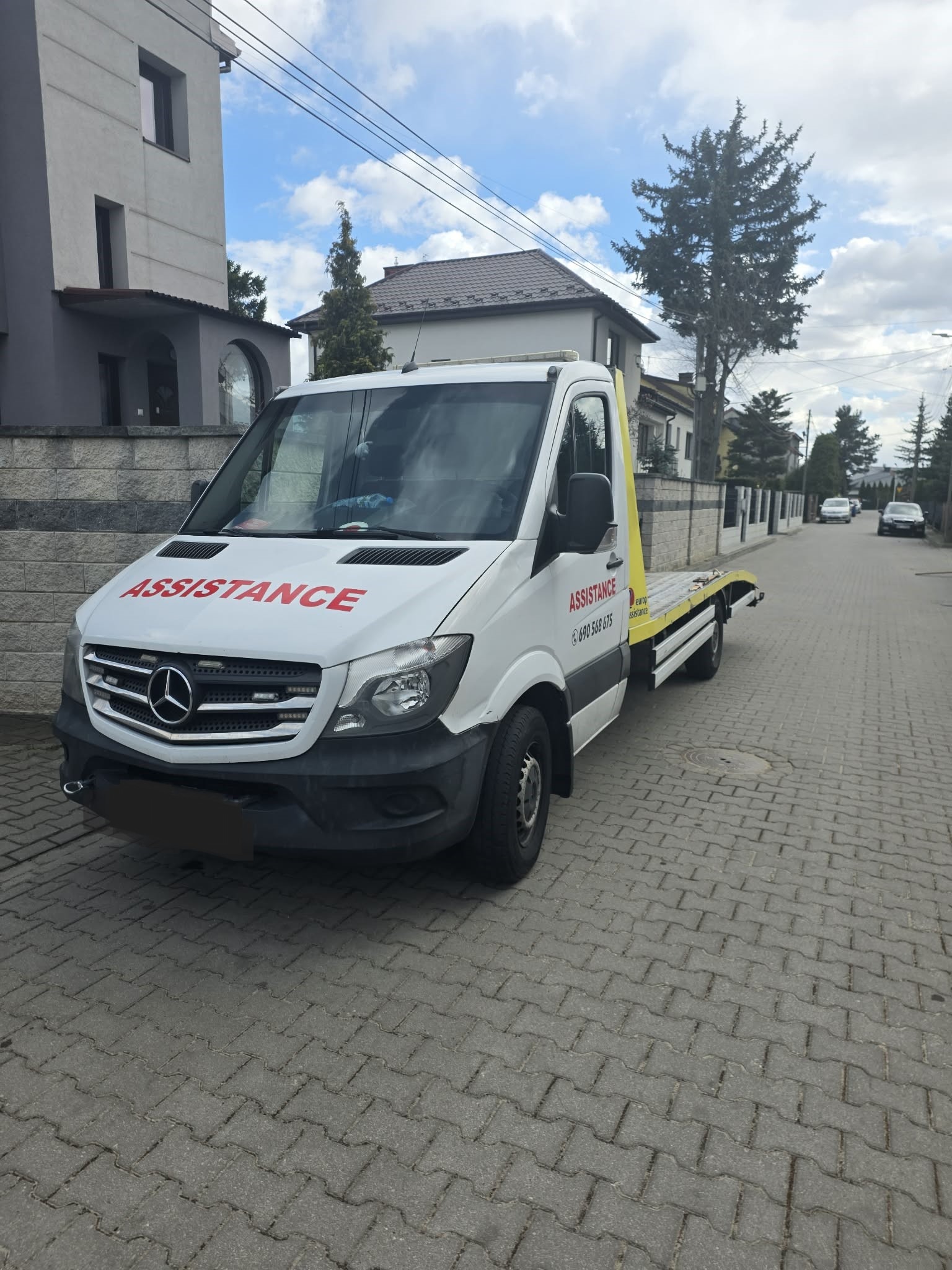 Biała laweta Mercedes Assistance na brukowanej ulicy, z widocznym numerem telefonu i napisem 'Assistance' na masce. W tle domy i drzewa.