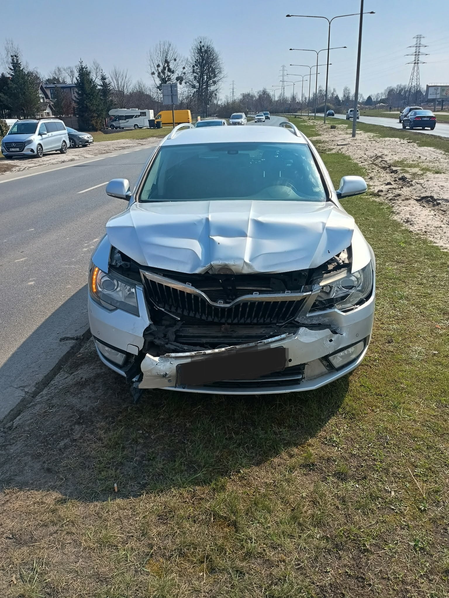 Srebrna Skoda Superb kombi z uszkodzonym przodem po kolizji, widoczna na poboczu drogi. Zniszczona maska, grill i reflektor. W tle droga i zabudowania.