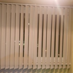 Bianex Jacek Łakomiec - Białe plisy pionowe zamontowane w oknie, widoczne z wnętrza pomieszczenia o kremowych ścianach. Częściowo widoczny mebel z jasnego drewna.