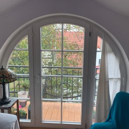 Bianex Jacek Łakomiec - Białe, łukowe okno z widokiem na balkon i zieleń, oświetlone lampą Tiffany. Wnętrze pokoju z elementami dekoracyjnymi i meblami. Charakterystyczny kształt okna.