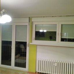 Bianex Jacek Łakomiec - Białe okna z plisami w pokoju z grzejnikiem i lampą sufitową. Widoczne trzy okna: jedno potrójne balkonowe i dwa mniejsze, pojedyncze. Ściany w odcieniach bieli i limonki.