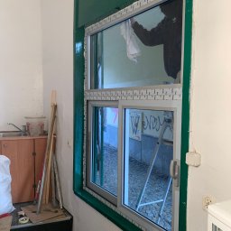 Bianex Jacek Łakomiec - Montaż okna PCV z zieloną ramą. Osoba czyści szybę od wewnątrz. Widoczne narzędzia i drabina. Ujęcie z boku, wnętrze pomieszczenia w trakcie prac wykończeniowych.