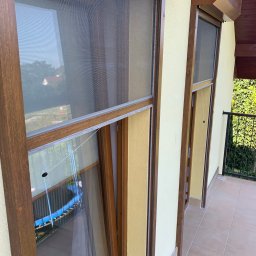 Bianex Jacek Łakomiec - Dwa okna z brązowymi ramami i moskitierami na balkonie, widok z zewnątrz. Na szybach odbija się niebieski basen. Żółta elewacja budynku.