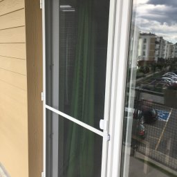 Bianex Jacek Łakomiec - Biała rama okienna z moskitierą na tle budynku mieszkalnego i parkingu. Widoczne detale mocowania moskitiery i odbicie otoczenia w szybie.