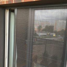 Bianex Jacek Łakomiec - Zamknięta, szara moskitiera plisowana na oknie balkonowym, z widokiem na budynki mieszkalne w tle. Widoczny fragment drewnianego ogrodzenia balkonu.