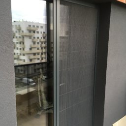 Bianex Jacek Łakomiec - Szara plisa okienna zamontowana w oknie balkonowym, widok z bliska. W tle budynek mieszkalny odbijający się w szybie. Minimalistyczny design, nowoczesne wykończenie.