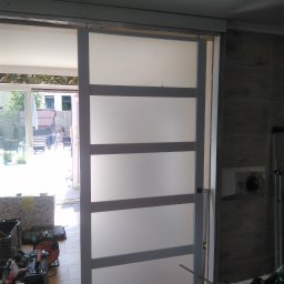 Bianex Jacek Łakomiec - Zamontowane drzwi przesuwne z czterema matowymi panelami, widoczne narzędzia i fragment wnętrza z wyjściem na zewnątrz. Ujęcie z bliska, z widocznymi detalami montażu.