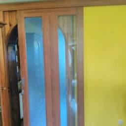 Bianex Jacek Łakomiec - Drewniane drzwi przesuwne z dwoma szklanymi panelami, widoczne na tle żółtej ściany i fragmentu drewnianej boazerii. Widoczny fragment wnętrza za drzwiami.