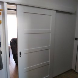 Bianex Jacek Łakomiec - Białe, przesuwne drzwi z czterema panelami, częściowo otwarte, ukazujące fragment wnętrza z drewnianą podłogą i osobą w środku. Minimalistyczny design, jasne oświetlenie.