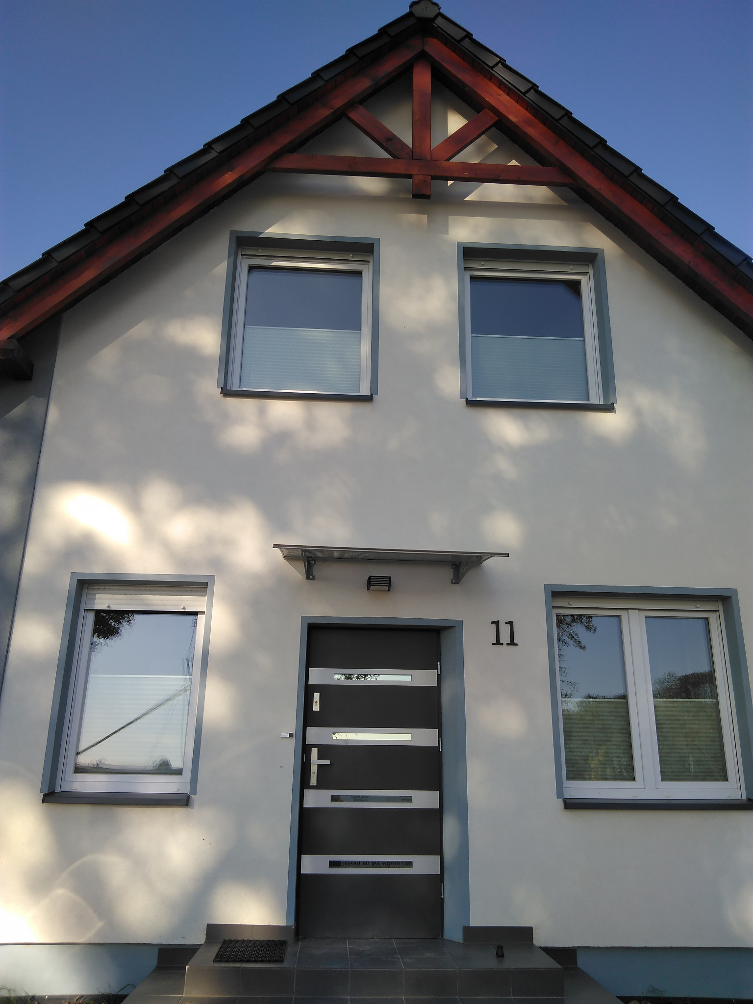 Elewacja domu z nowoczesnymi oknami z plisami i drzwiami wejściowymi w kolorze antracytowym. Widoczne elementy drewniane konstrukcji dachu i numer domu.