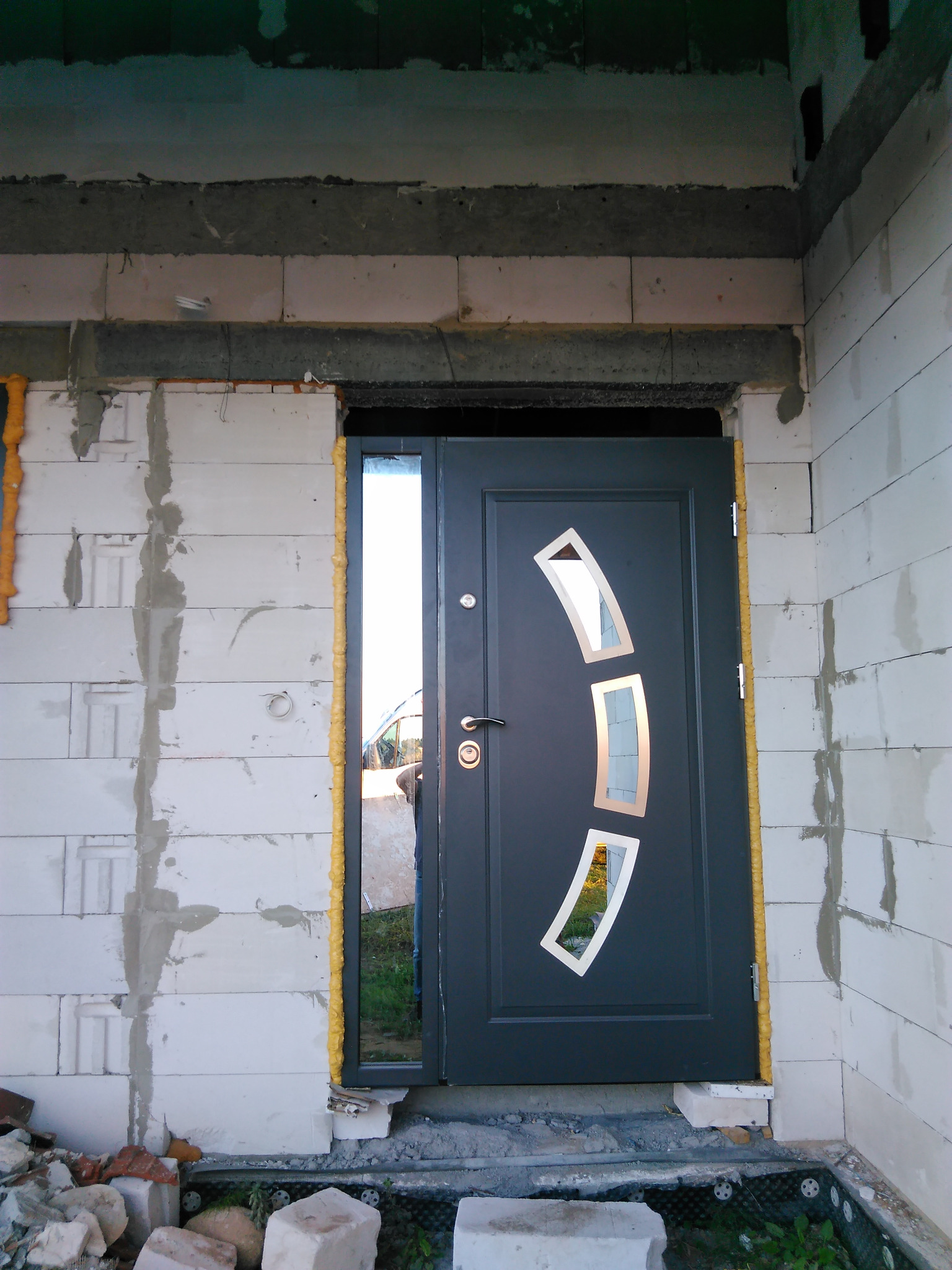 Montaż drzwi wejściowych z szybą boczną w surowym stanie. Widoczne pianowanie ościeżnicy i niedokończone ściany z betonu komórkowego. Drzwi w kolorze grafitowym z dekoracyjnymi szybami.