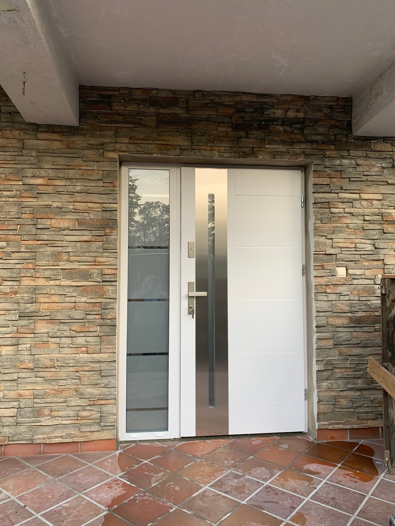 Białe drzwi wejściowe z przeszkleniem i stalową wstawką, osadzone w kamiennej elewacji. Widoczna terakota na podłodze i fragment rusztowania.