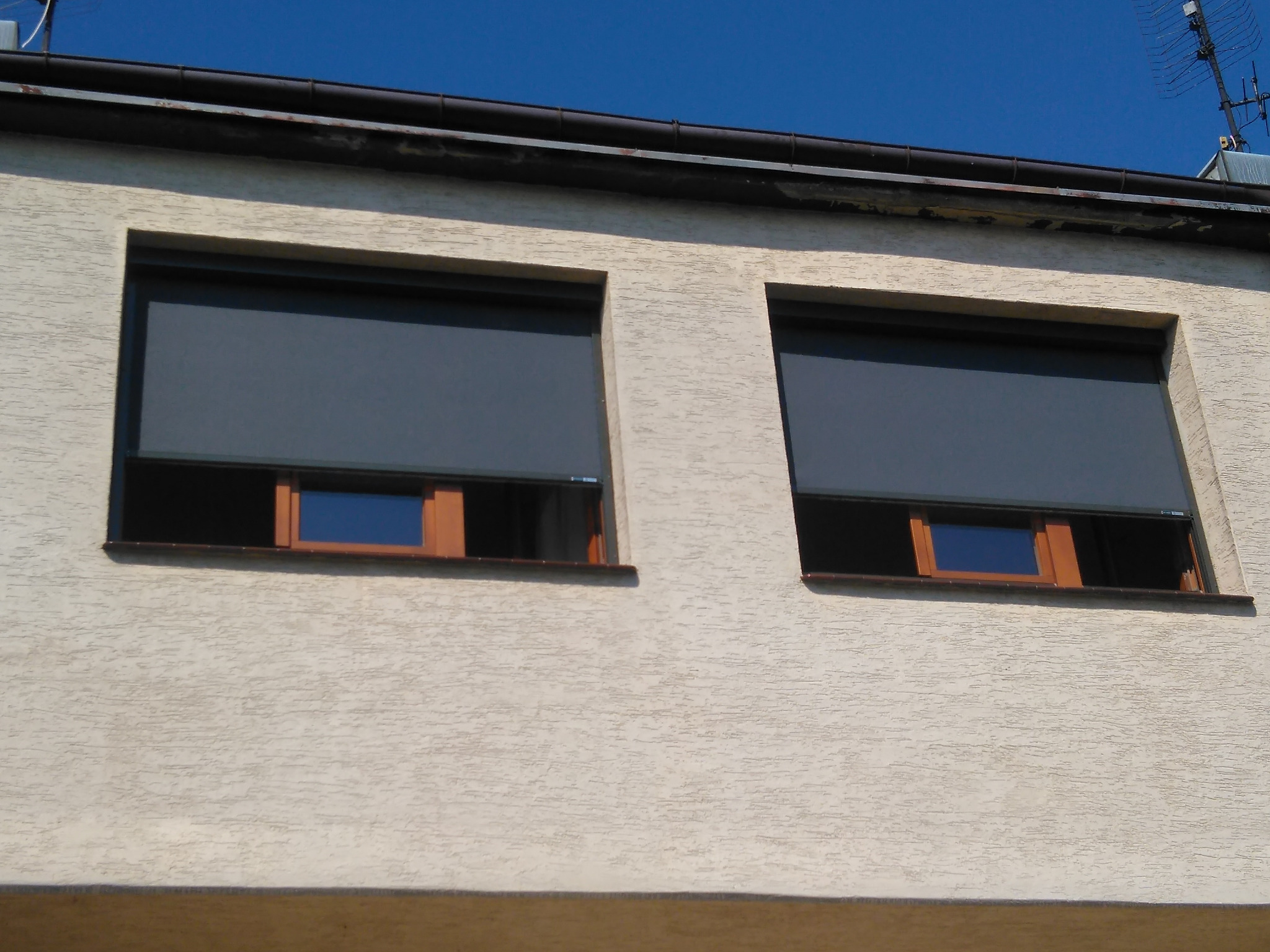 Dwa okna z drewnianą ramą, zasłonięte szarymi plisami, na tle jasnej, teksturowanej elewacji budynku w słoneczny dzień. Widoczny fragment dachu i anteny.