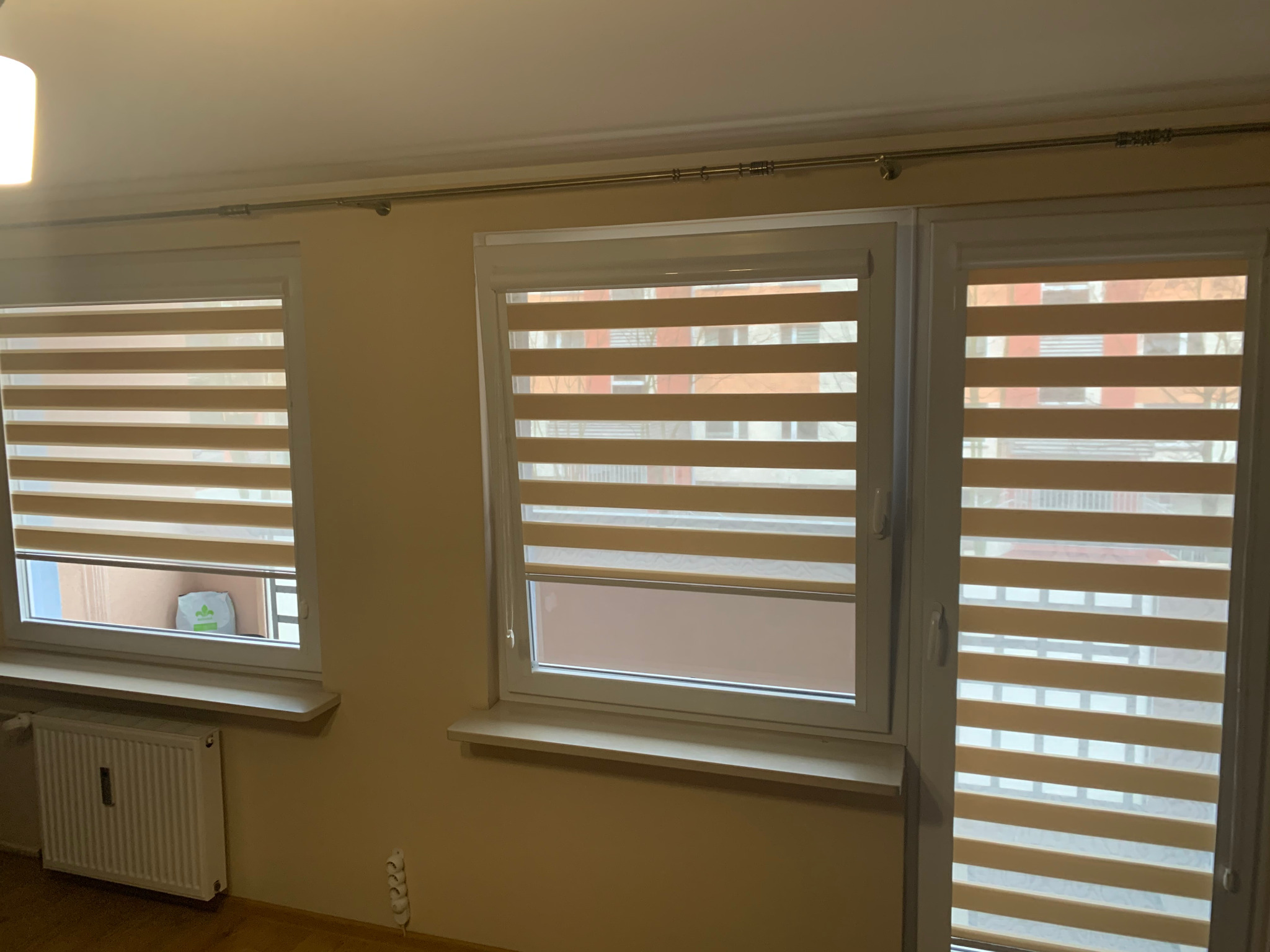 Trzy okna z zamontowanymi plisami okiennymi typu dzień i noc w kolorze beżowym. Widok na zewnątrz przez okna. Na parapecie leży torba z zakupami.