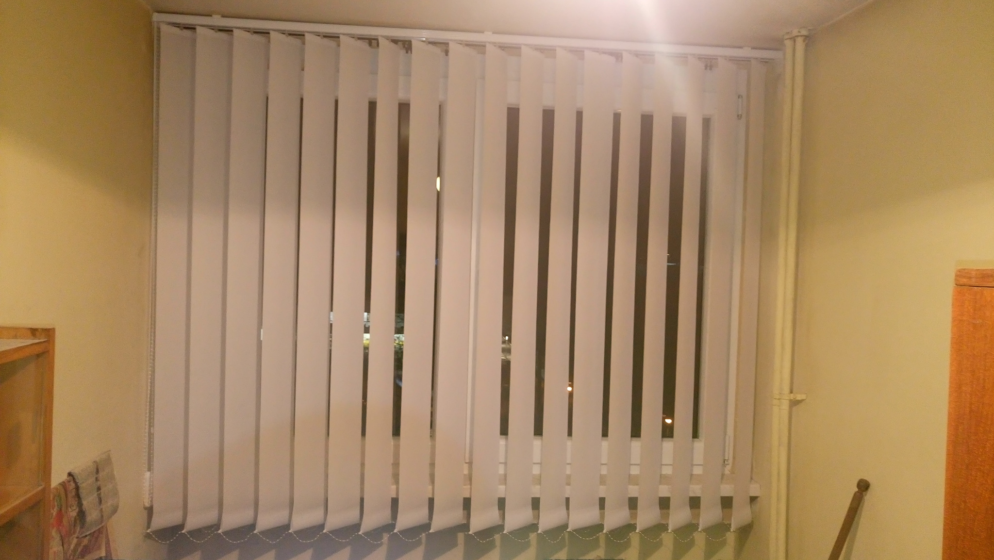 Białe plisy pionowe zamontowane w oknie, widoczne z wnętrza pomieszczenia o kremowych ścianach. Częściowo widoczny mebel z jasnego drewna.