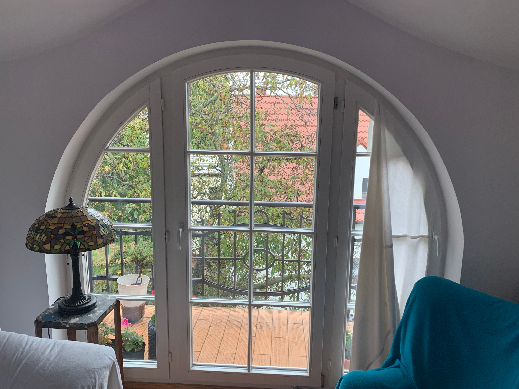 Białe, łukowe okno z widokiem na balkon i zieleń, oświetlone lampą Tiffany. Wnętrze pokoju z elementami dekoracyjnymi i meblami. Charakterystyczny kształt okna.